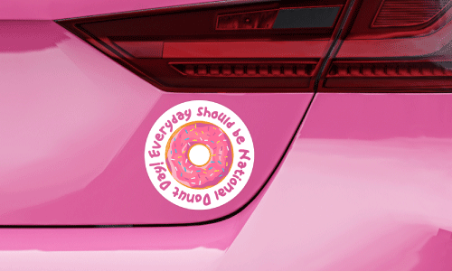 Back bumper of pink sedan. There is a round bumper sticker of a pink sprinkled donut. Pink text on white in a circular shape says Every Day Should Be National Donut Day.